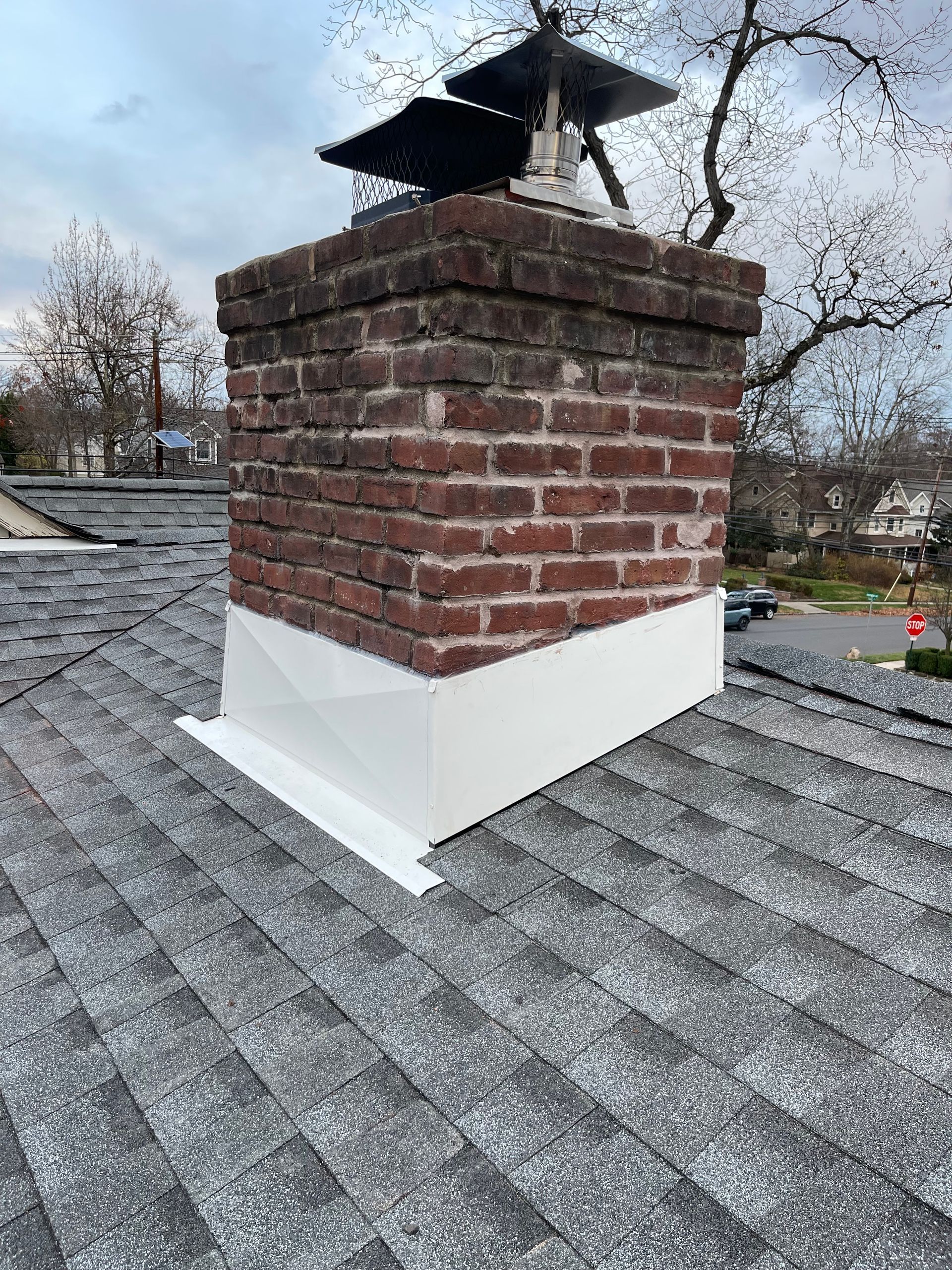 Brick chimney on a gray shingled roof with a black metal cap and white flashing.