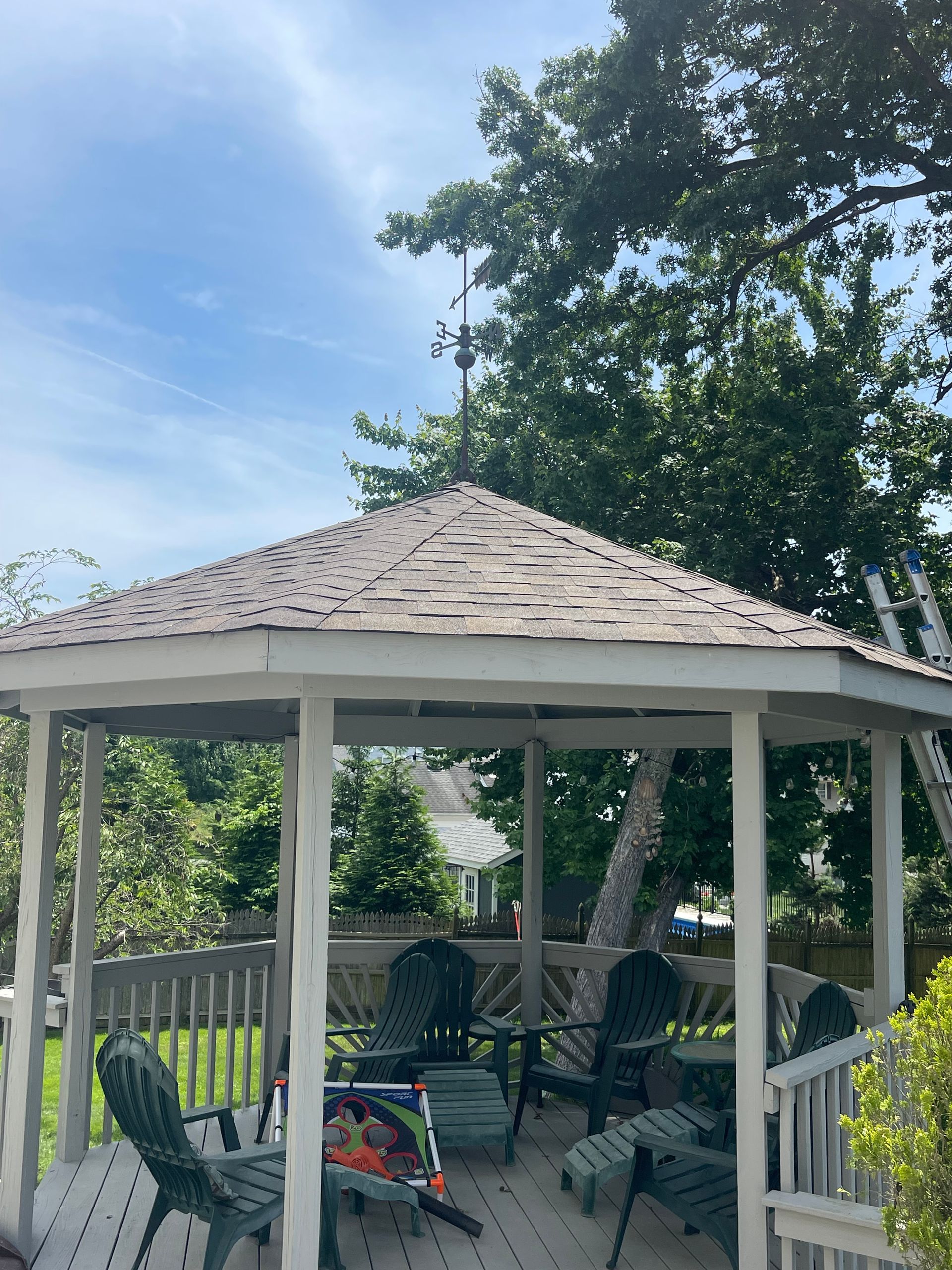 Backyard gazebo with chairs and a bicycle under a shingled roof, surrounded by trees and a fence.