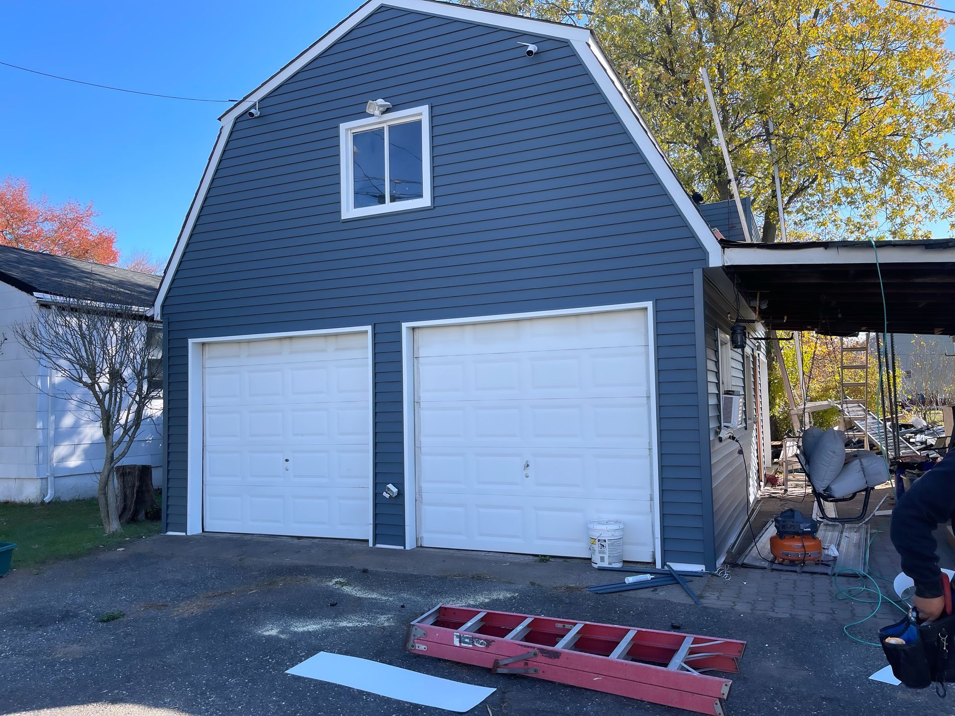 Gray barn-style garage with two white doors, a red ladder on the ground, and a person at right.