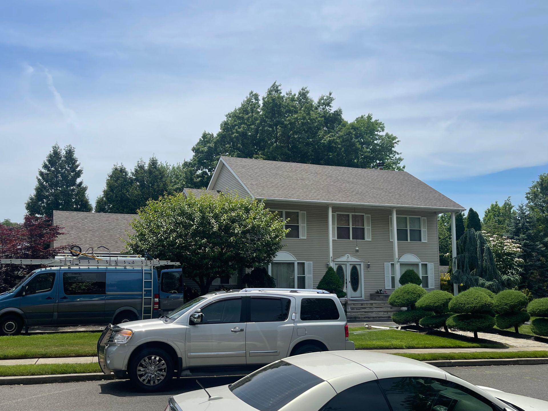 Suburban house with two parked vehicles on a sunny street, trees and shrubs in front