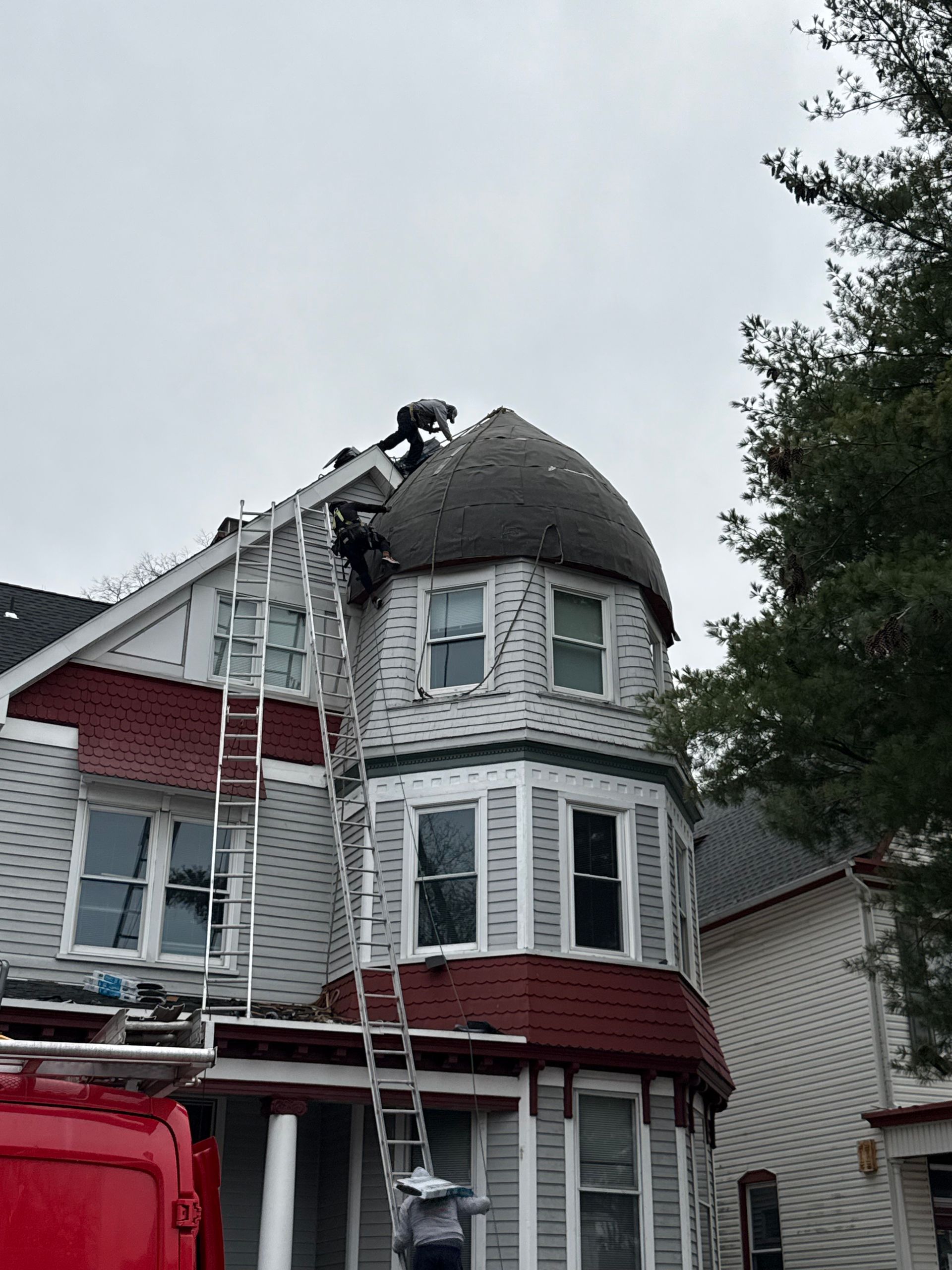 Firefighters on a ladder at a gray Victorian house with a damaged roof and smoke visible near the top.