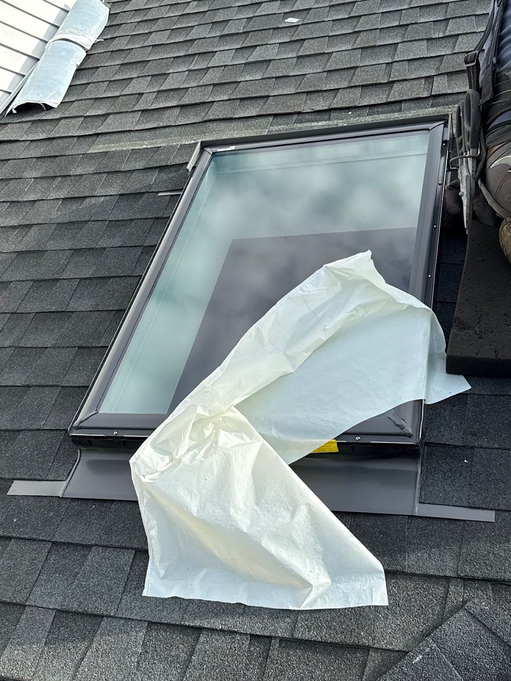 Partially open skylight on a shingled roof with a white plastic sheet draped over it