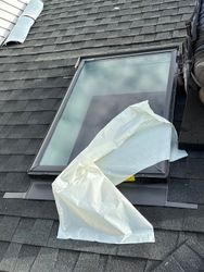 Partially open skylight on a shingled roof with a white plastic sheet draped over it