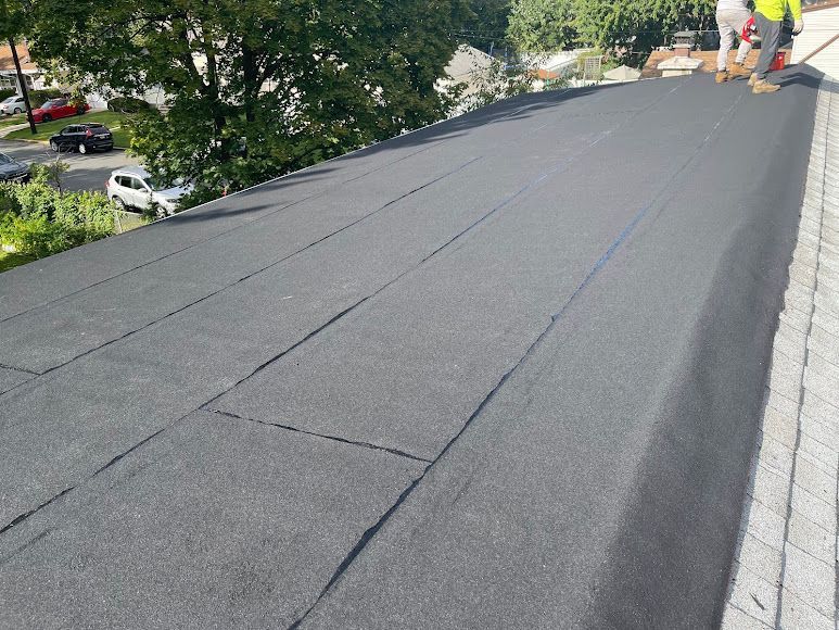 Workers installing black roofing felt on a sloped roof near trees and houses