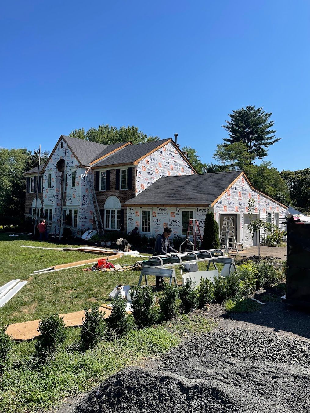 Stone house with gray roof and patio, surrounded by landscaped yard under a clear blue sky.