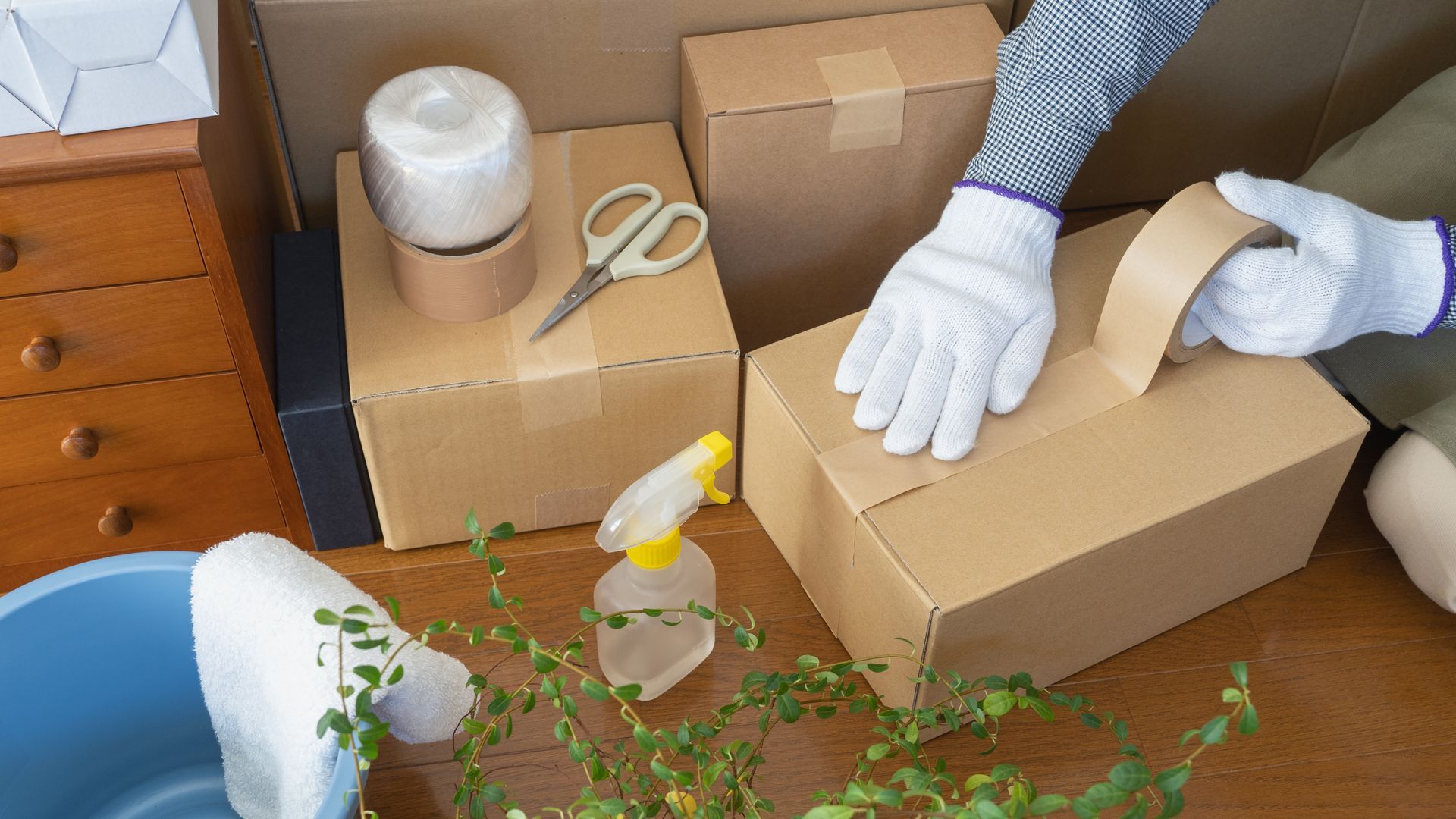 Person sealing a cardboard box with tape, preparing for moving; various boxes, tools visible.