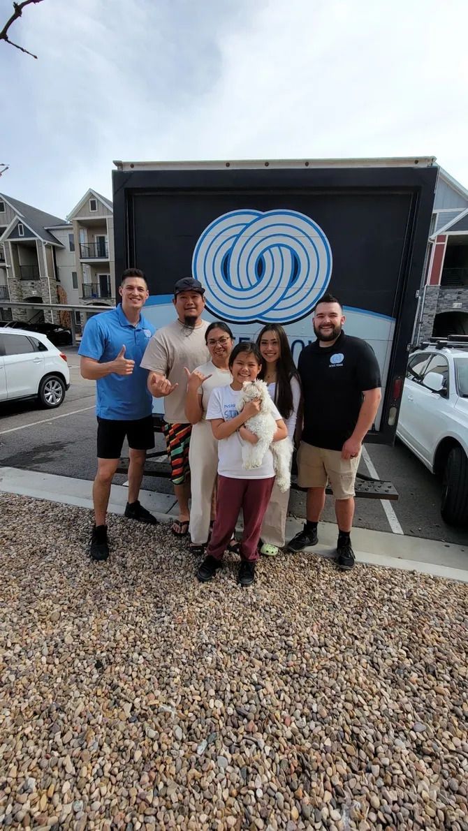 Group of people posing in front of a moving truck with a blue logo, outside an apartment building.