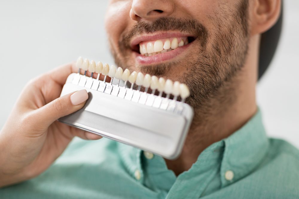 A Man Is Holding A Tooth Colour Chart In Front Of His Mouth — Mittagong Dental Care In Mittagong, NSW