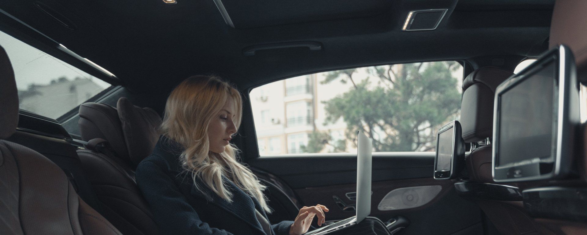 A woman is sitting in the back seat of a car using a laptop computer.