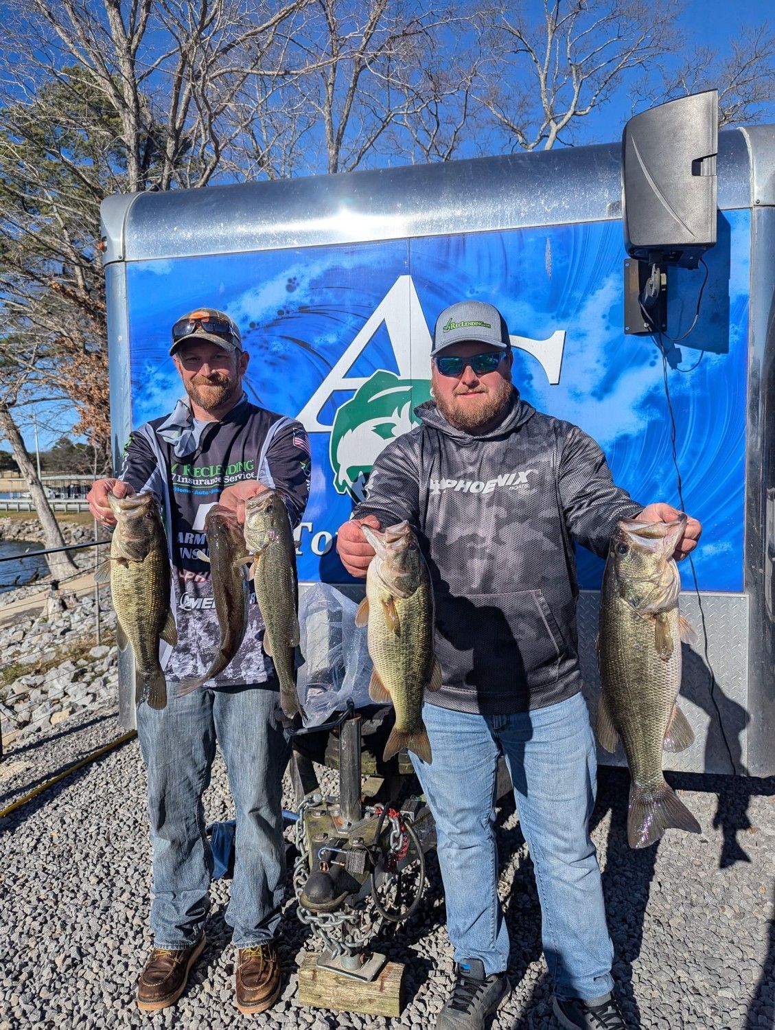 Two anglers holding several large fish in front of a blue wall outdoors, standing on gravel.