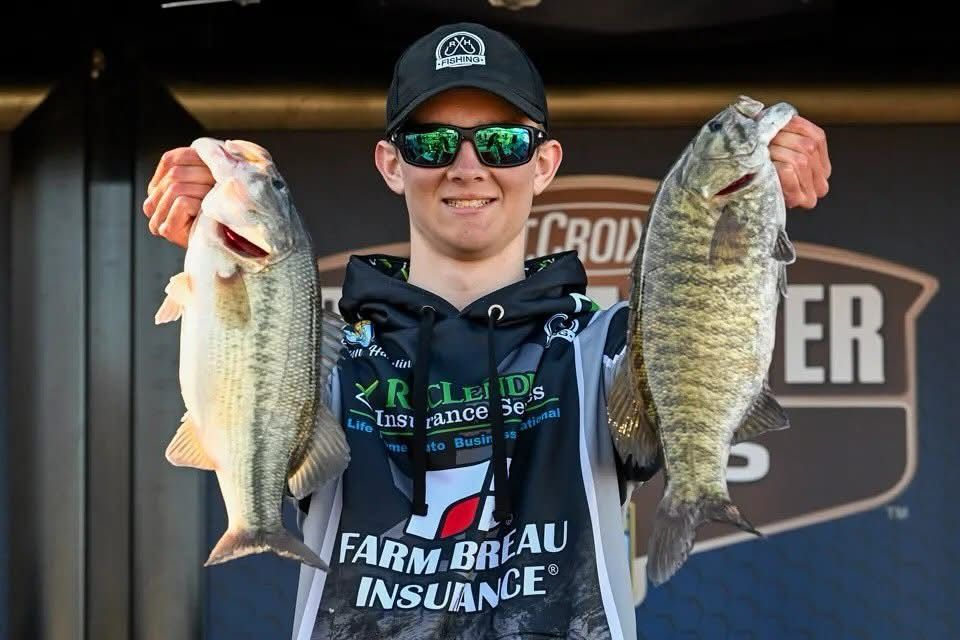 Angler holding two large fish on a stage, wearing a cap, sunglasses, and a black jersey