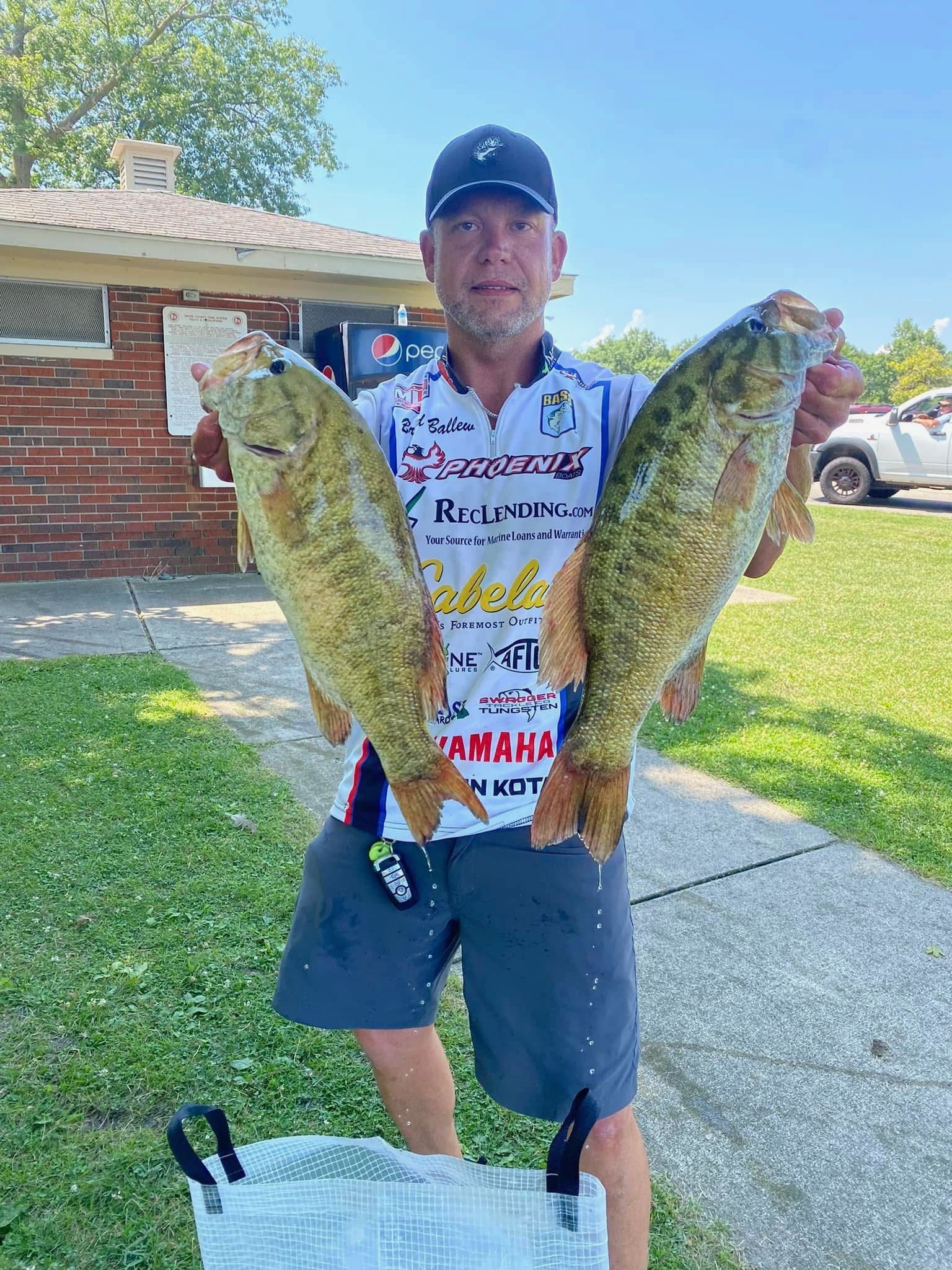 Angler holding two large fish outdoors on a sunny driveway, wearing a fishing jersey and cap.