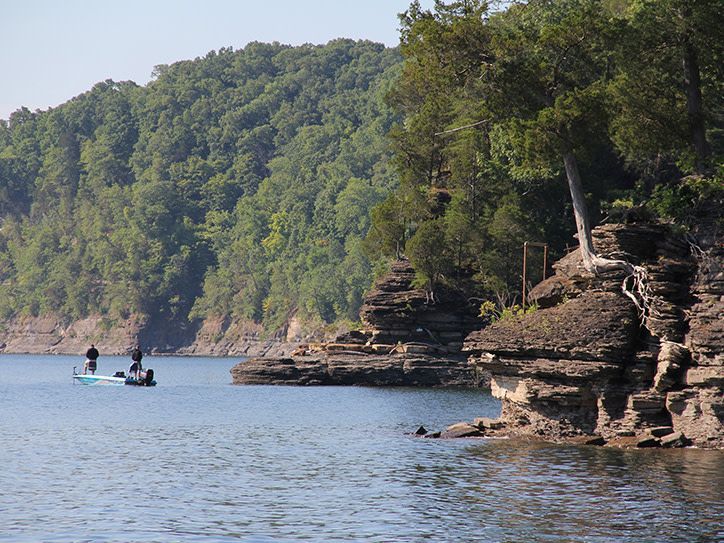 Two fishermen on a calm blue lake beside rocky cliffs and a forested shoreline