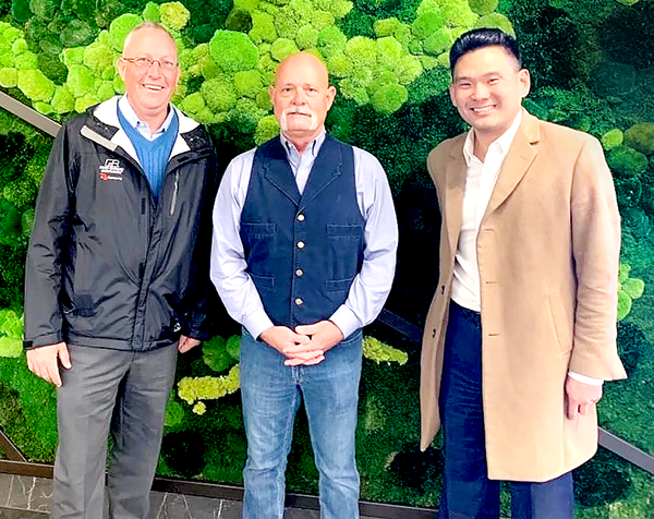 Three men standing in front of a green leafy wall, smiling at the camera.
