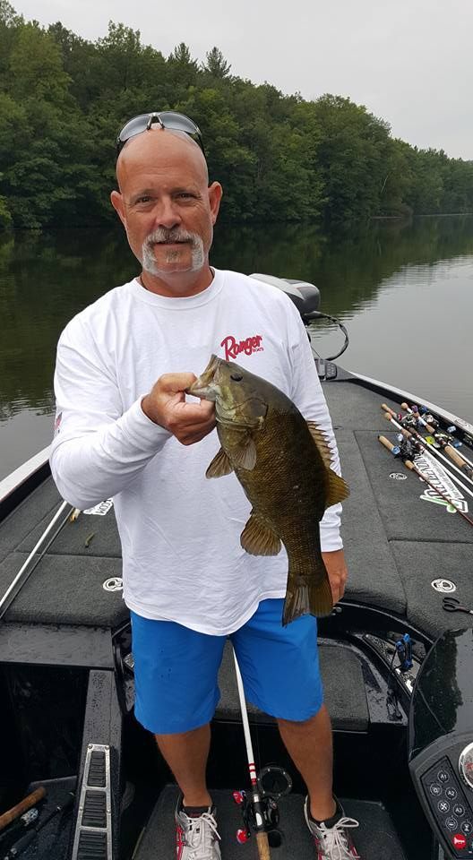 Man on a boat holding a large fish, with fishing rods and a calm, wooded shoreline behind him.