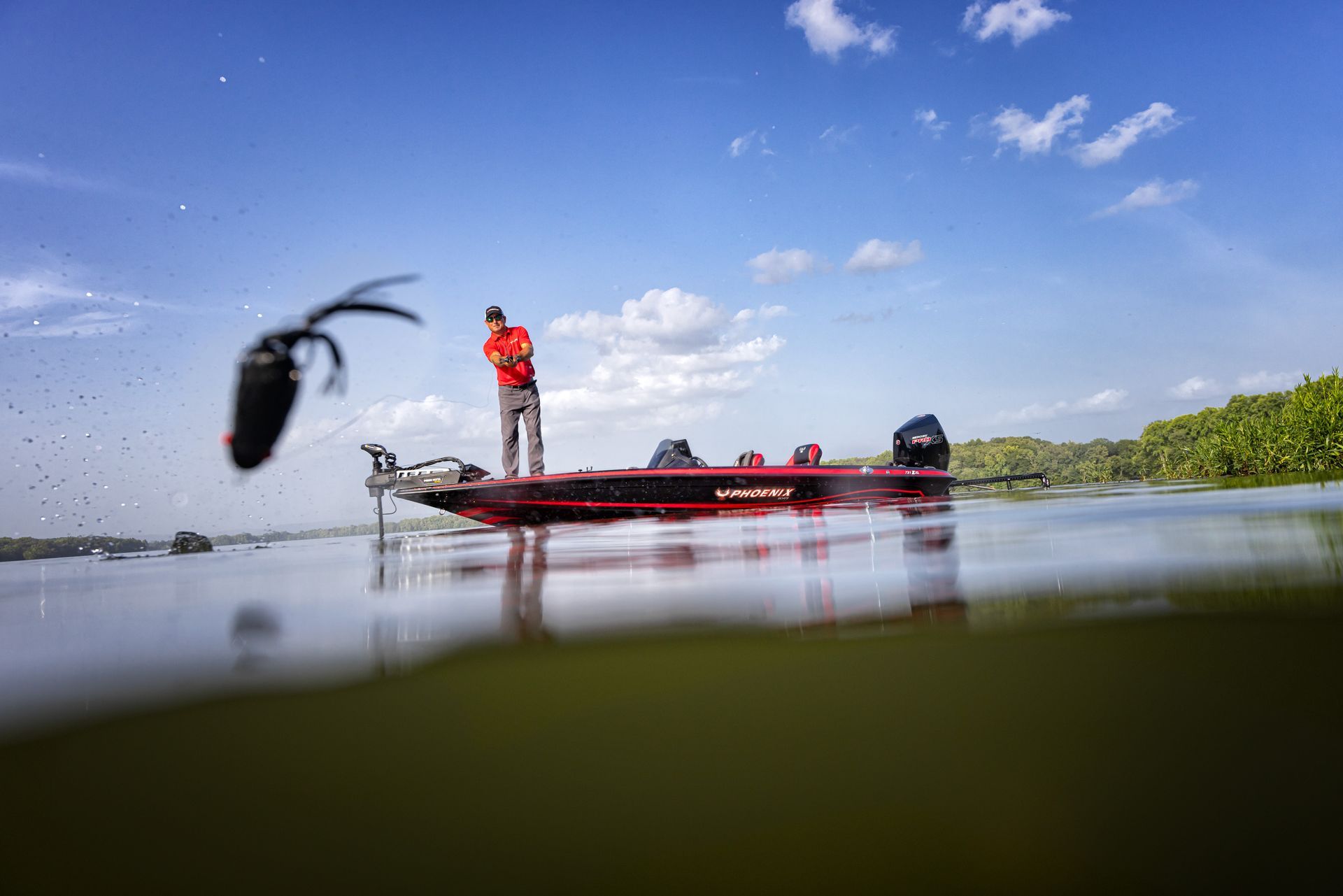 Angler standing on a fishing boat on calm water under a blue sky, with a lure splashing in the foreground