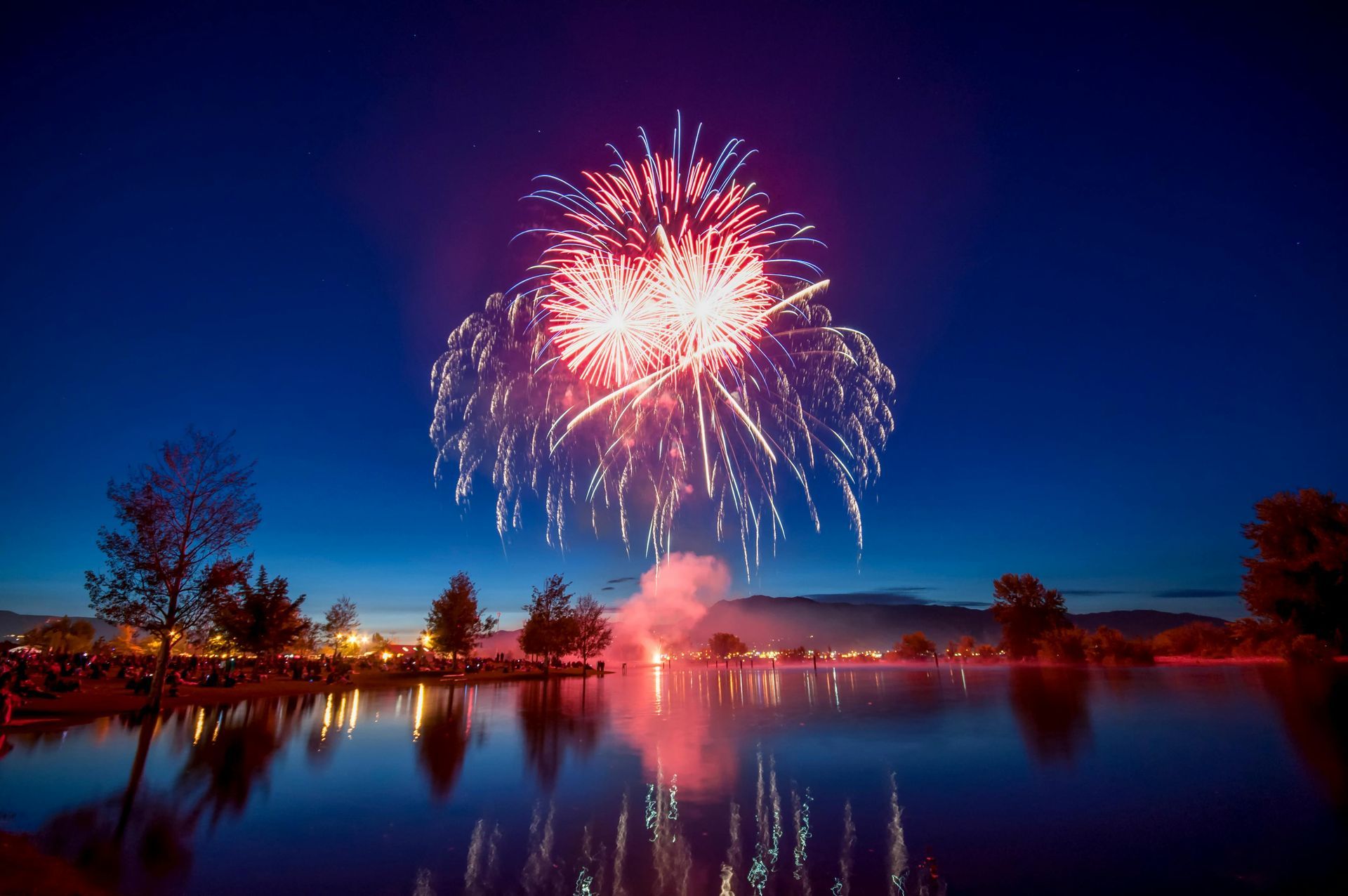 Fireworks burst over a calm lake at dusk, with colorful reflections on the water.
