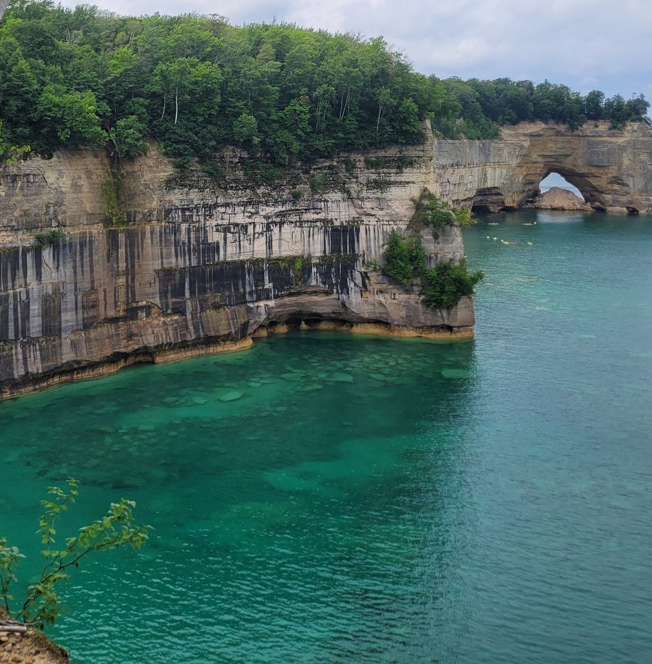 Turquoise water below a rocky cliff topped with green trees and an arched sea cave.