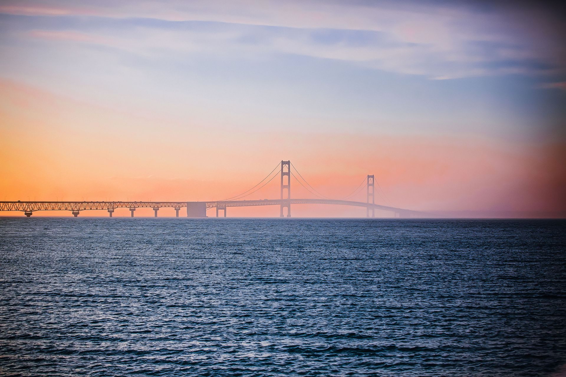 The Mackinac suspension bridge over calm water at sunset, with pink-orange sky and dark blue sea.
