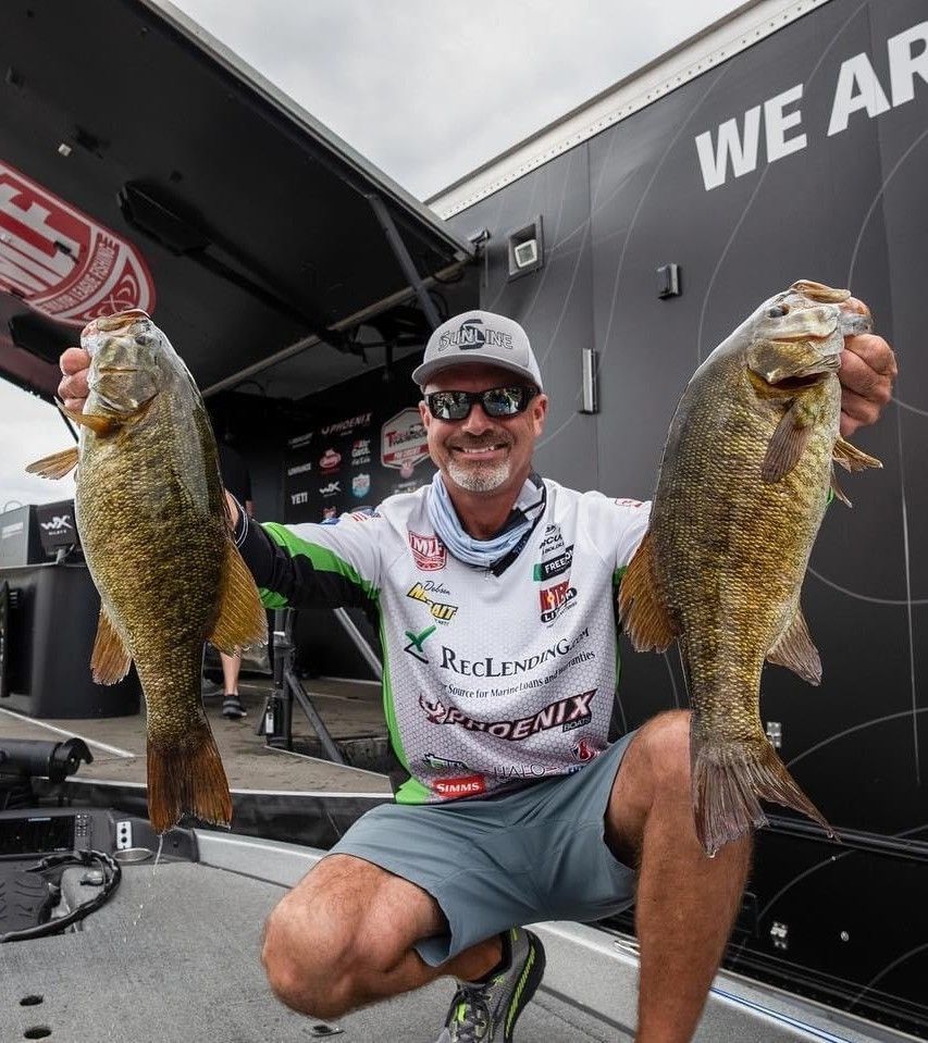 Man holding two large fish, smiling outdoors beside a black truck at a fishing event