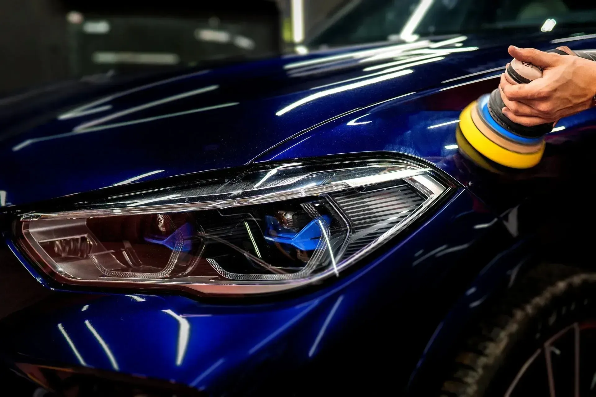 Hand polishing the headlight and hood of a dark blue car with a cloth and yellow-blue wristbands