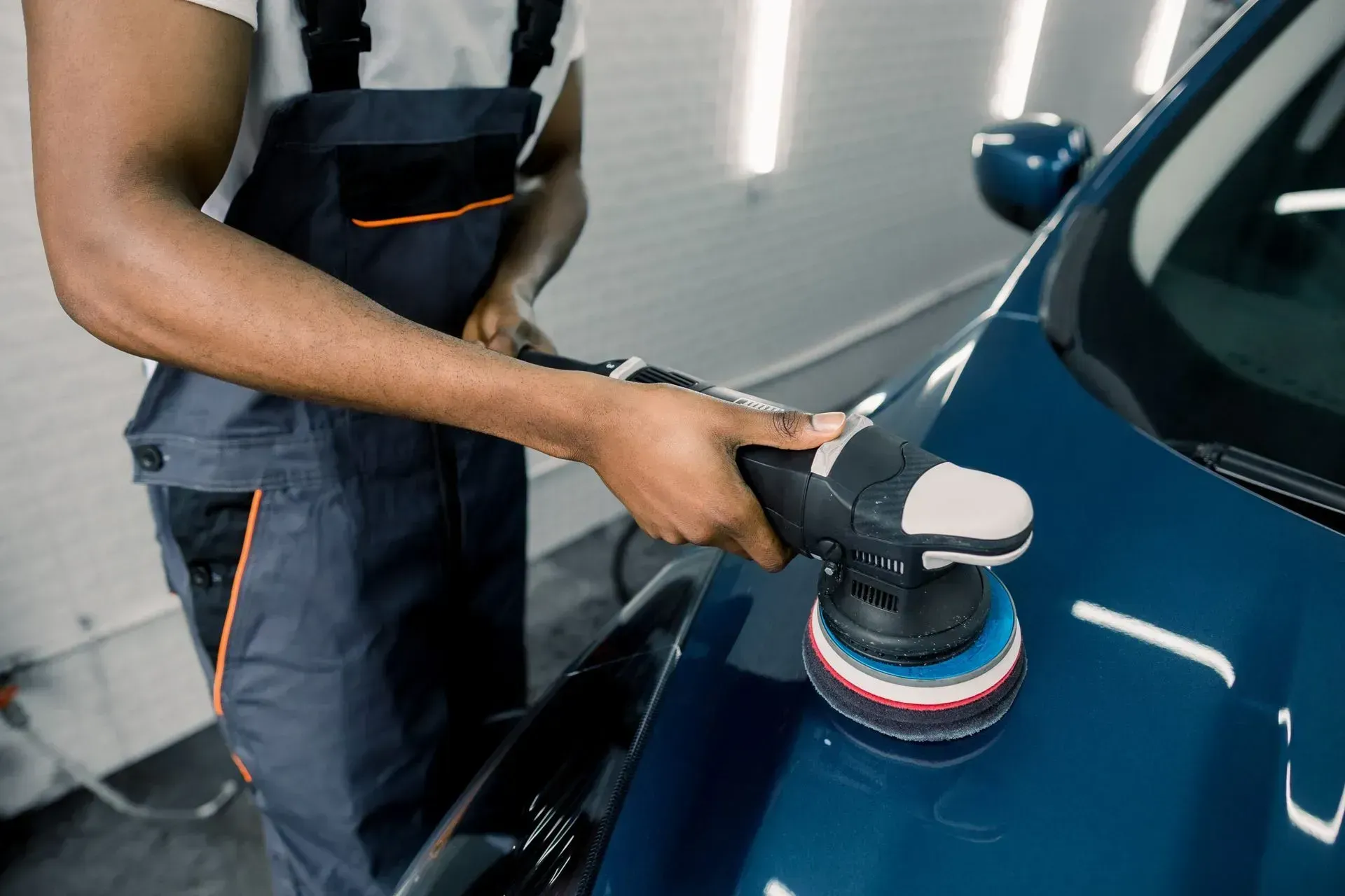 Person polishing a blue car with a handheld buffer in a garage