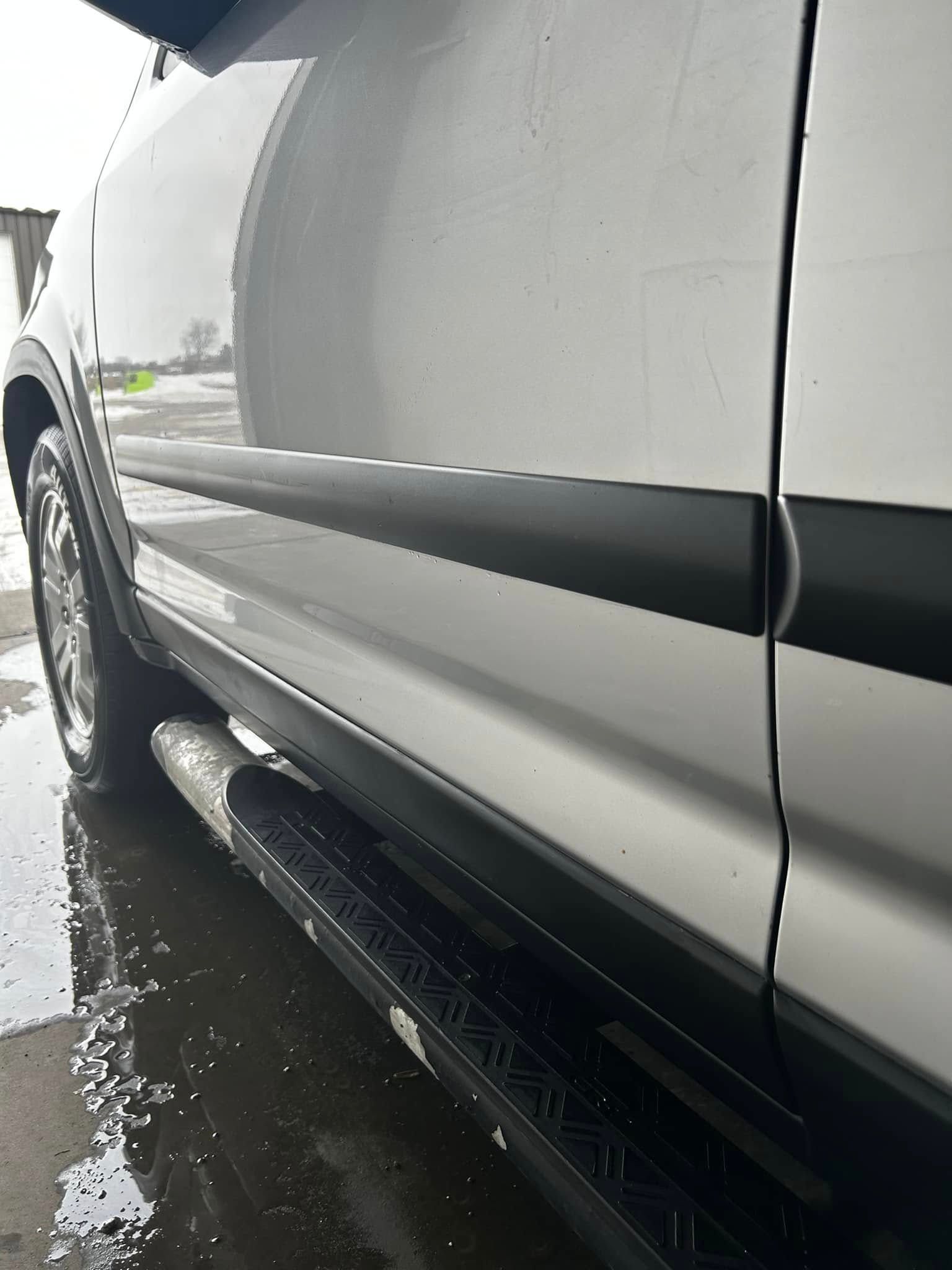 Close-up of a white SUV’s side and black running board, wet from rain on a driveway