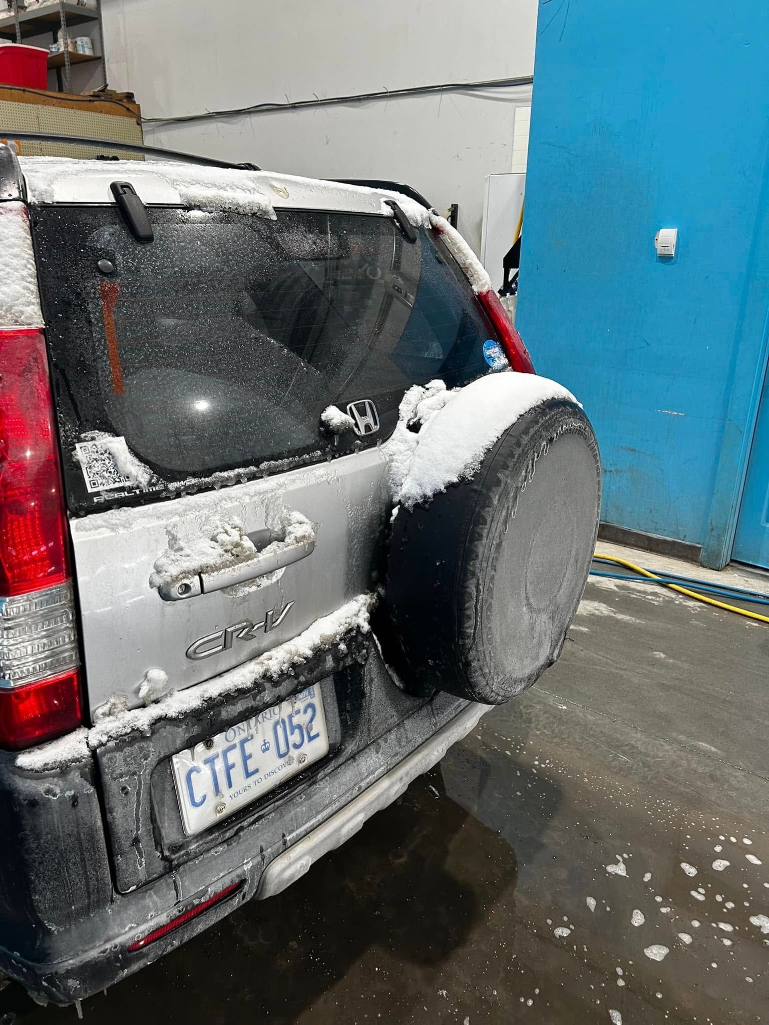Snow-covered black SUV parked by a blue wall, rear view with spare tire on the back.