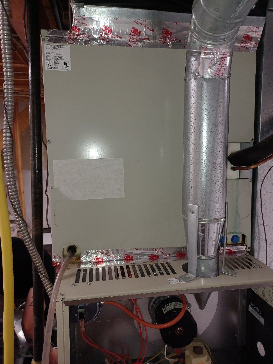 Furnace unit with ductwork in a basement setting. Gray metal, silver duct, orange wires visible.