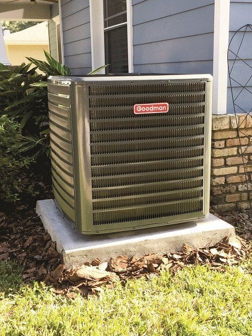 Goodman air conditioning unit on a concrete pad near a brick wall and light blue house.