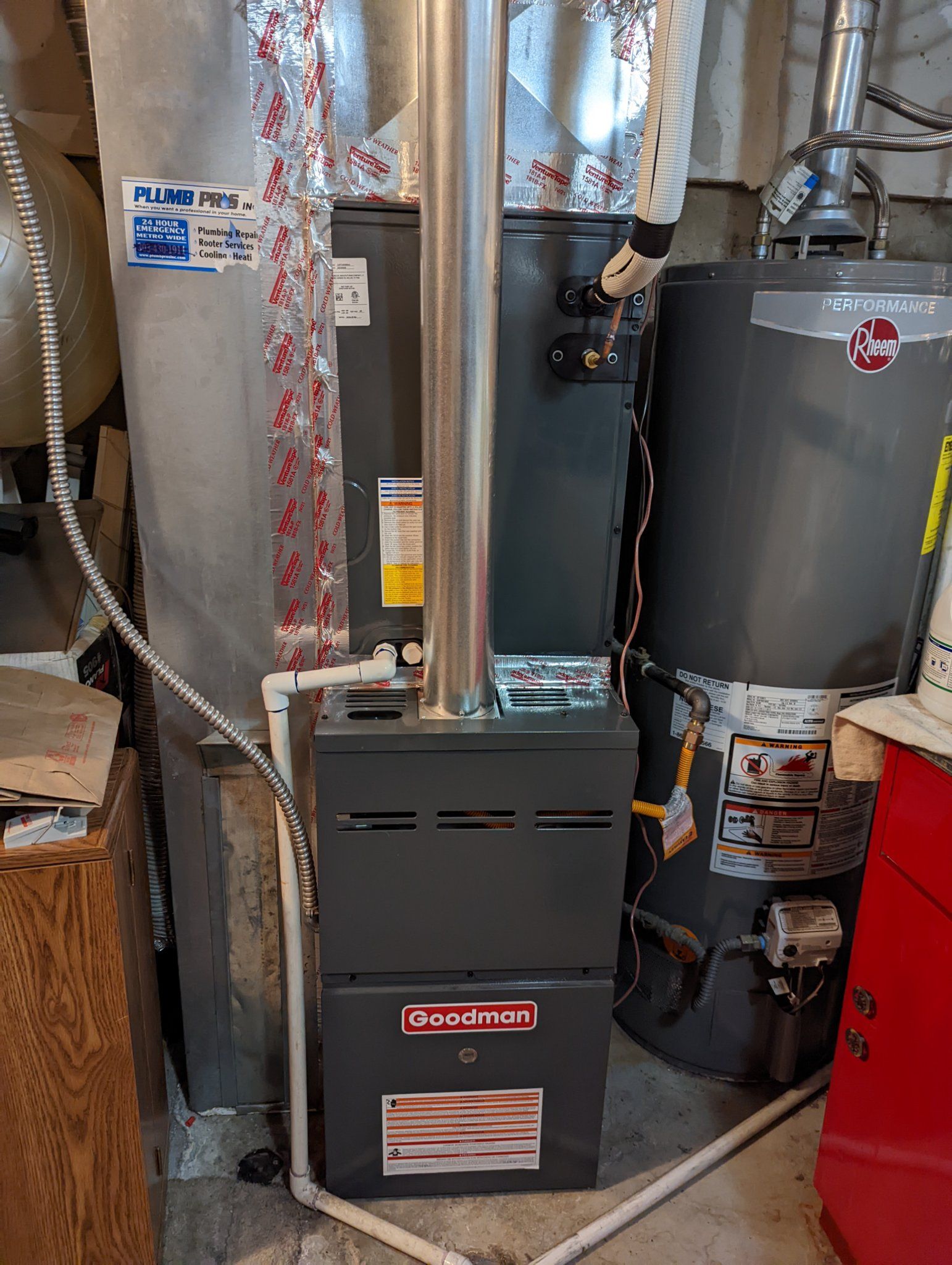 Furnace and water heater in a basement. Goodman furnace is dark grey with a tall silver exhaust pipe.