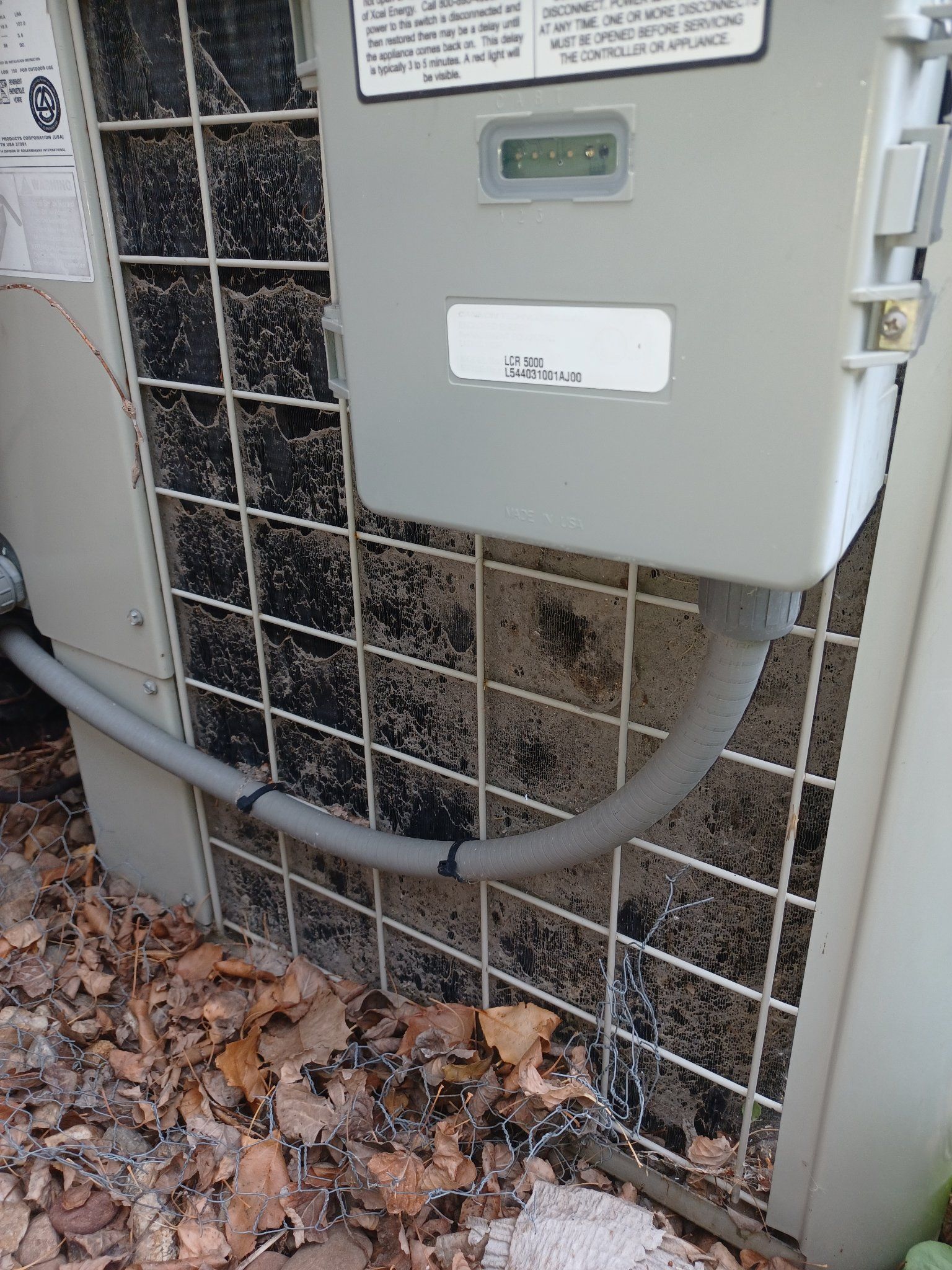 Air conditioning unit with electrical box and protective grid. Grey and brown tones, outdoors with leaves.