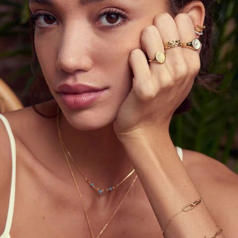 Close-up portrait of a woman wearing gold jewelry, resting her face on her hand against a blurred green background