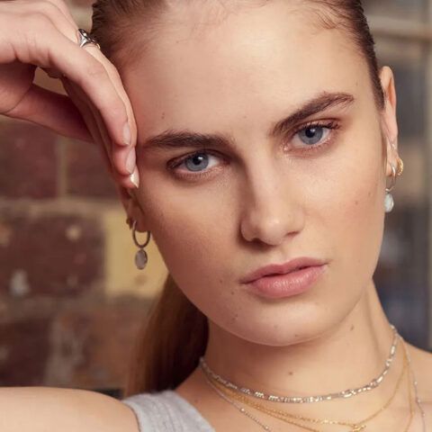 Close-up portrait of a woman with blue eyes, resting her hand on her temple, wearing delicate gold jewelry