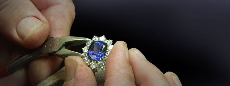 Hands use pliers to hold a blue gemstone in a silver ring for repair or setting.