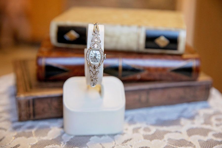 Gold watch with diamond-encrusted band displayed upright on a white cushion, with books blurred in the background