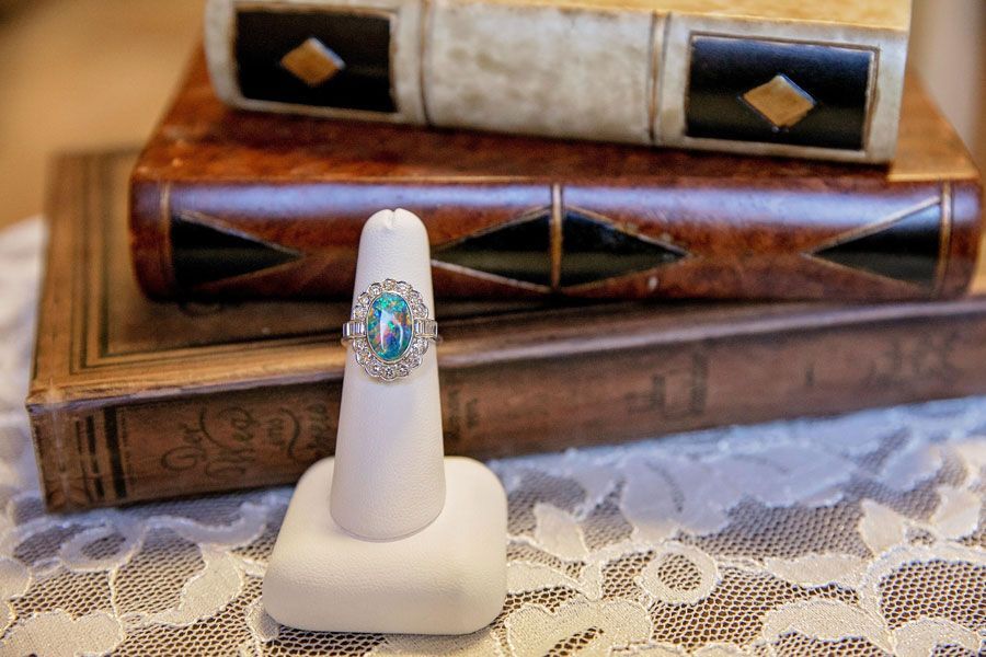 White ring stand stand with silver-blue gem surrounded by diamonds, in front of carved wooden boxes on lace tablecloth