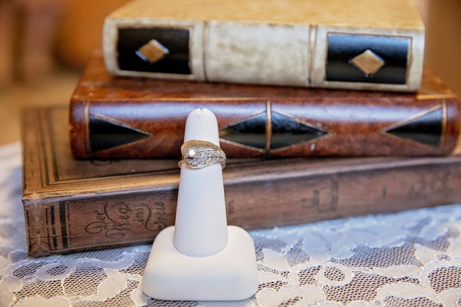 Diamond ring on white ring stand in front of stacked decorative wooden boxes on a lace tablecloth.