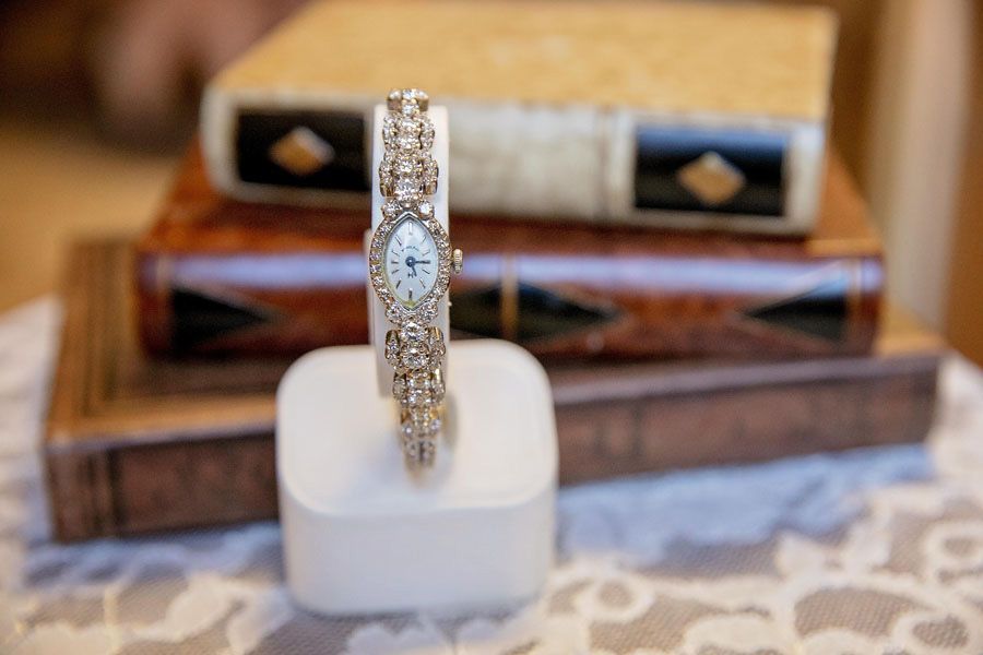 Gold watch with diamond-encrusted band displayed upright on a white cushion, with books blurred in the background