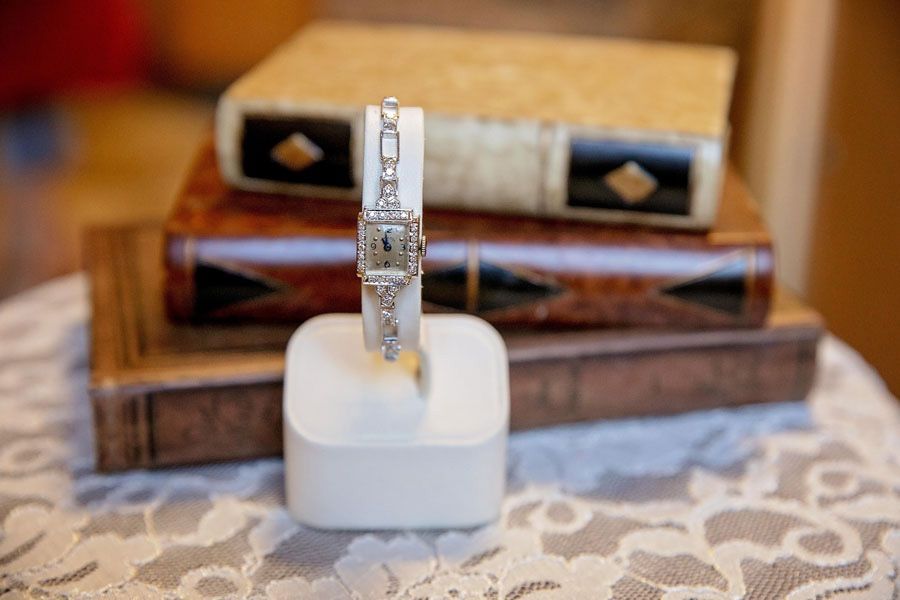 Gold watch with diamond-encrusted band displayed upright on a white cushion, with books blurred in the background