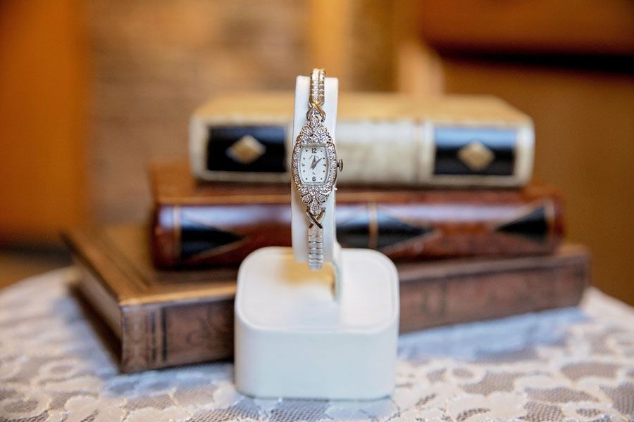 Gold watch with diamond-encrusted band displayed upright on a white cushion, with books blurred in the background
