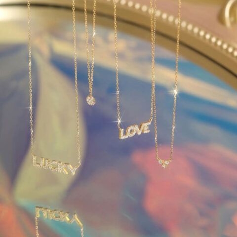 Gold necklaces with hanging word charms spelling “lucky,” “love,” and “lucky” against a pastel background