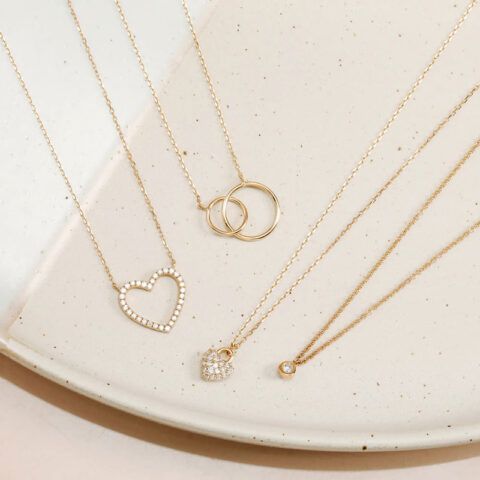 Gold necklaces with heart, ring, and solitaire pendants on a cream surface