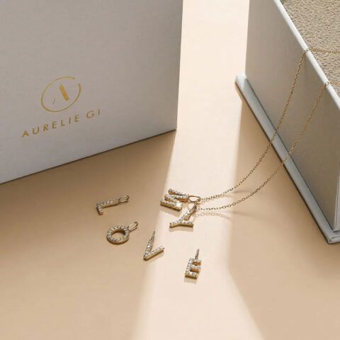 Gold jewelry pieces and a necklace displayed on a beige surface beside a white box labeled “Aurelie Gi.”