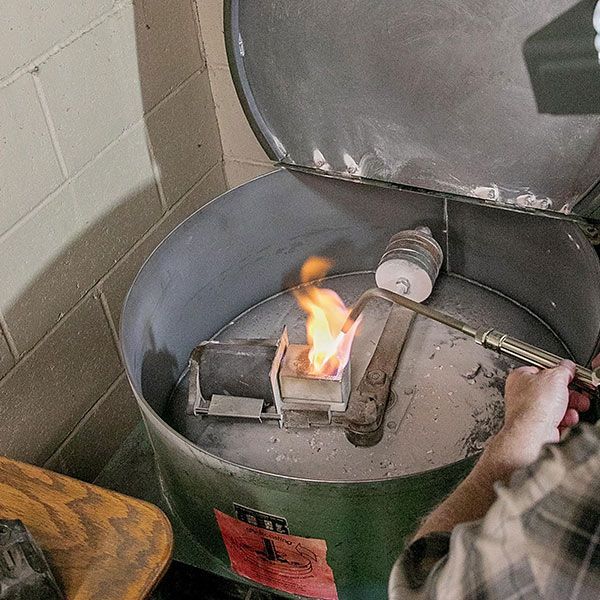 Green metal burner with a small flame inside, next to a tiled wall in a workshop-like room