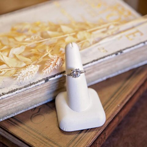 Diamond engagement ring on a white jewelry stand beside a book on a wooden surface