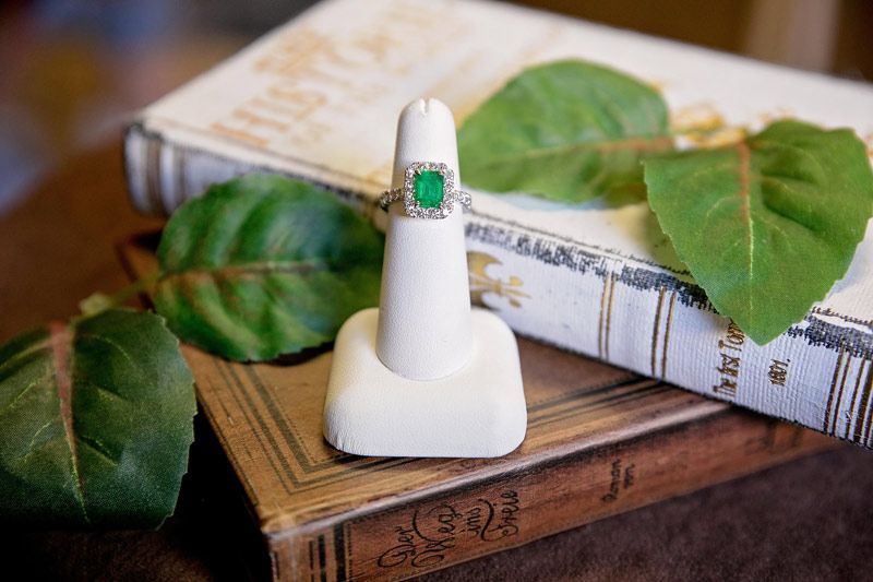 White ring with green stone on a stand beside leaves and a stacked book