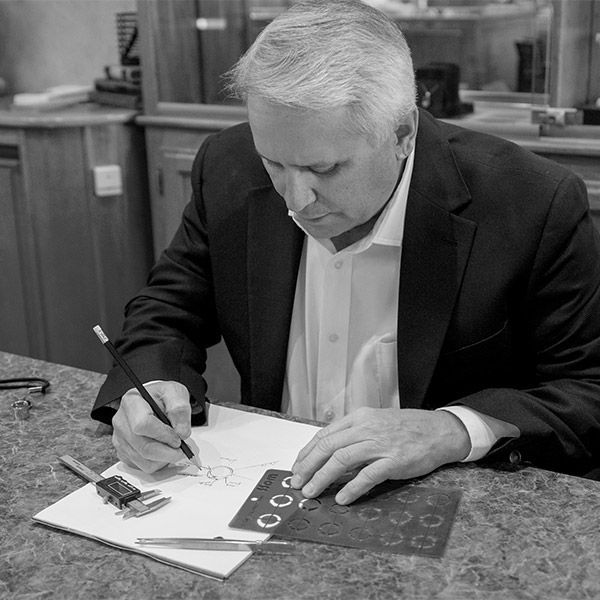 Man in a dark blazer sketches at a table, focused on a drawing with drafting tools nearby.