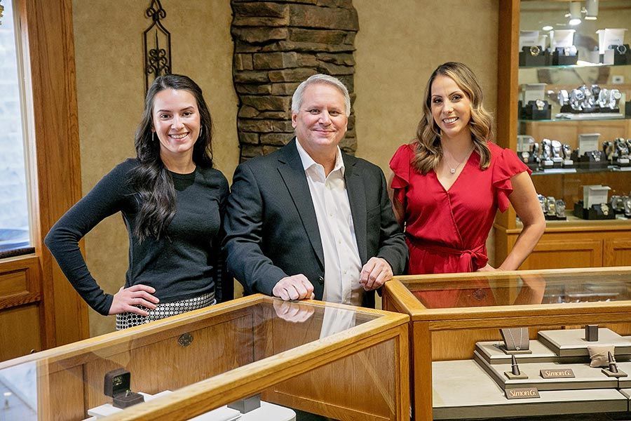 Three people standing behind a glass display case in a jewelry store, smiling at the camera.