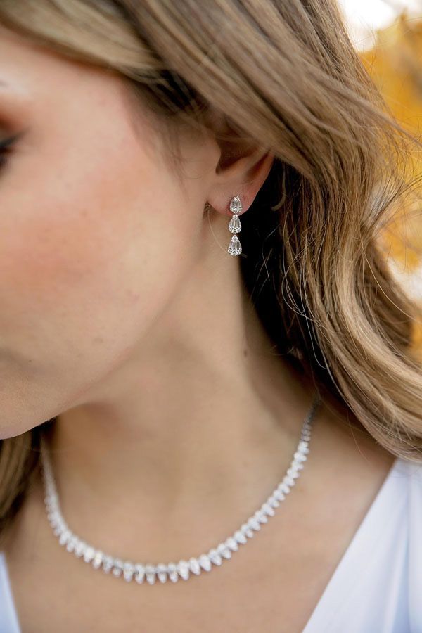 Close-up of a woman wearing a pearl necklace and dangling earrings against a blurred autumn background